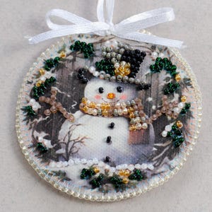 Peut inclure: Une d&eacute;coration ronde en tissu blanc avec un motif de bonhomme de neige. Le bonhomme de neige porte un chapeau haut de forme noir avec des perles dor&eacute;es et vertes, une &eacute;charpe brune avec des perles dor&eacute;es et blanches, et a des yeux noirs et un nez orange. La d&eacute;coration est entour&eacute;e de perles vertes et blanches.
