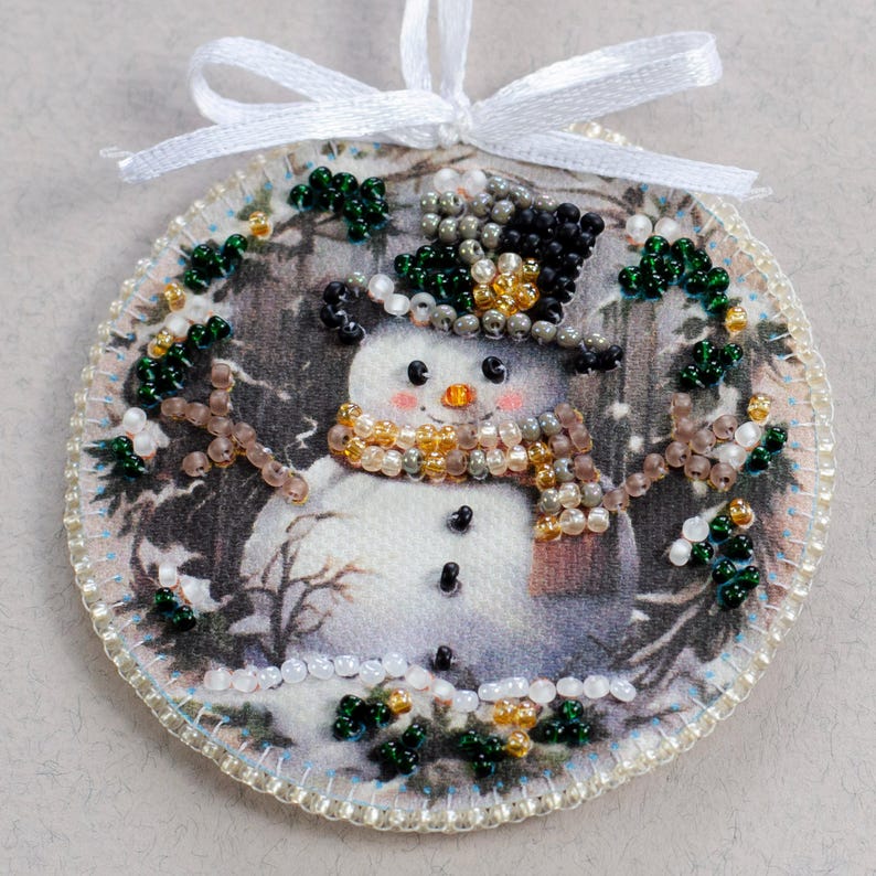 Peut inclure: Une d&eacute;coration ronde en tissu blanc avec un motif de bonhomme de neige. Le bonhomme de neige porte un chapeau haut de forme noir avec des perles dor&eacute;es et vertes, une &eacute;charpe brune avec des perles dor&eacute;es et blanches, et a des yeux noirs et un nez orange. La d&eacute;coration est entour&eacute;e de perles vertes et blanches.