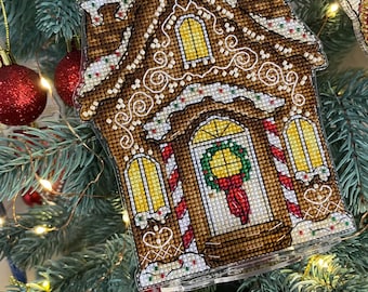 May include: A gingerbread house Christmas ornament with white icing and red candy canes. The house has a wreath on the door and a window with yellow light.