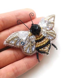 May include: A detailed bee brooch, hand-stitched with beads. The wings are white with silver and gold detailing, and the body is striped with black and gold beads. A black bead forms the head, with wire antennae.