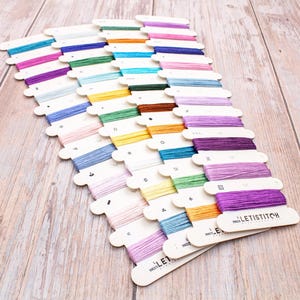 May include: A collection of colorful embroidery floss spools arranged on a light wood surface. The spools are in various shades of blue, pink, green, yellow, orange, and purple. Each spool is labeled with a number and the brand name "LETISTITCH".