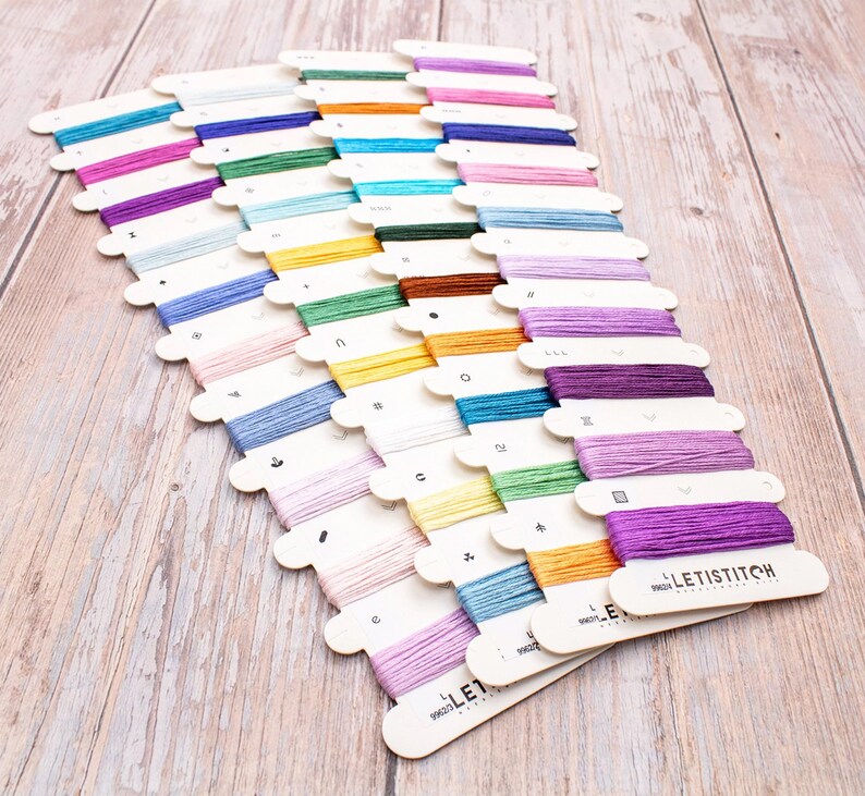May include: A collection of colorful embroidery floss spools arranged on a light wood surface. The spools are in various shades of blue, pink, green, yellow, orange, and purple. Each spool is labeled with a number and the brand name "LETISTITCH".