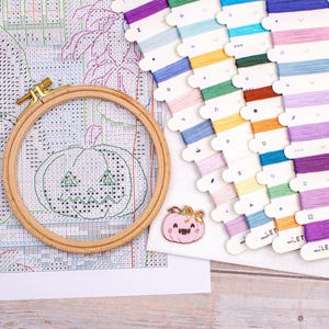 May include: A cross-stitch project in progress, featuring a wooden embroidery hoop with a stitched jack-o'-lantern design. Colorful embroidery floss in various shades is arranged nearby, along with a pink pumpkin pin. The project is on a white fabric.