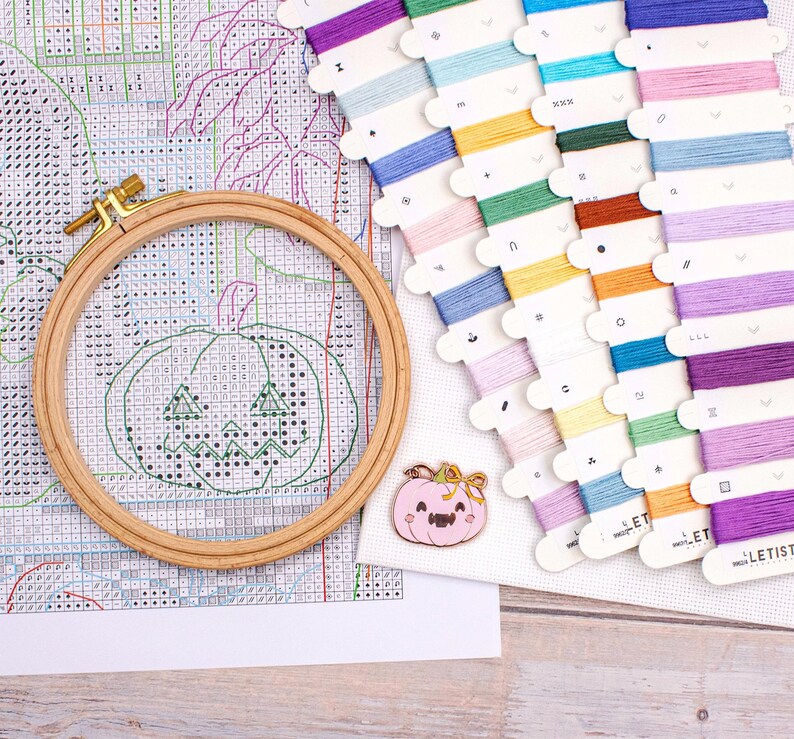 May include: A cross-stitch project in progress, featuring a wooden embroidery hoop with a stitched jack-o'-lantern design. Colorful embroidery floss in various shades is arranged nearby, along with a pink pumpkin pin. The project is on a white fabric.