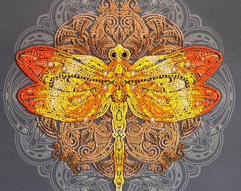 May include: A close-up of a beaded dragonfly with orange, yellow, and brown accents. The dragonfly is centered on a mandala-like design with intricate details.