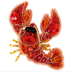 以下が含まれることがあります： 赤、オレンジ、金色のビーズと宝石を使った、手作りのロブスターブローチ。ロブスターの体は、さまざまなサイズの赤とオレンジのビーズで飾られ、黒いビーズの目と金のワイヤーのアクセントが施されています。