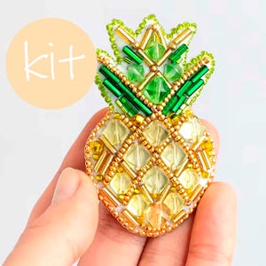 May include: A beaded pineapple brooch kit. The pineapple design features yellow, green, and gold beads. The word "kit" is in a light orange circle in the upper left corner. The brooch is held in a hand.