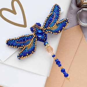 Puede incluir: Un broche de libélula azul y dorado con cuentas y un colgante de cristal. Las alas de la libélula están detalladas con cuentas azules y doradas, y el cuerpo está acentuado con una gran piedra preciosa azul.