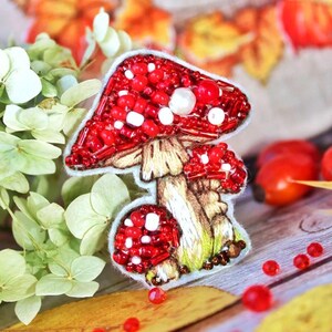 Op de afbeelding: Handgemaakte paddenstoelbroche met een rode, met kralen bezette hoed en witte stippen. De paddenstoel heeft een bruine steel en groene grasdetails aan de basis.