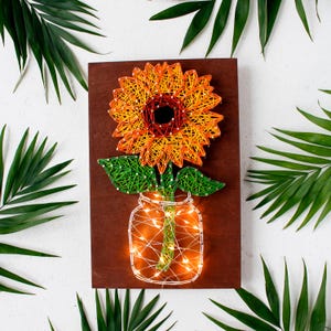 May include: A handcrafted string art piece featuring a sunflower in a mason jar. The sunflower is made with orange and yellow thread, the leaves are green, and the jar is illuminated with fairy lights. The artwork is mounted on a dark brown wooden board.