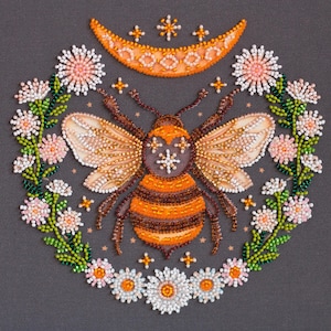 May include: A beaded embroidery design of a bee with orange and brown stripes, surrounded by a wreath of white and pink flowers. The bee has a crescent moon above it with stars.