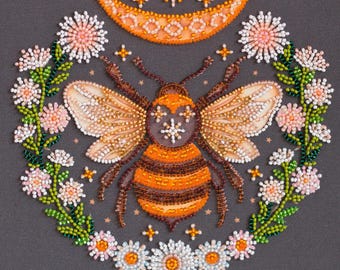 Könnte beinhalten: Eine Perlen-Stickerei mit einer Biene mit orange-braunen Streifen, umgeben von einem Kranz aus weißen und rosa Blüten. Die Biene hat einen Halbmond mit Sternen darüber.