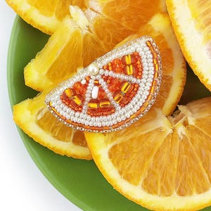 Könnte beinhalten: Eine Perlenbrosche in Form einer Orangenscheibe mit orangefarbenen, gelben und weißen Perlen. Die Brosche liegt auf einem Hintergrund aus frischen Orangenscheiben auf einem grünen Teller. Die Brosche hat einen Perlenakzent.