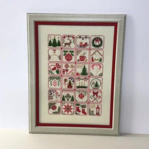May include: Framed cross-stitch artwork featuring a grid of Christmas-themed squares. Each square contains a different holiday image, including trees, reindeer, and ornaments. The frame is silver with a red inner border, creating a festive display.