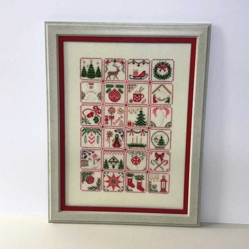 May include: Framed cross-stitch artwork featuring a grid of Christmas-themed squares. Each square contains a different holiday image, including trees, reindeer, and ornaments. The frame is silver with a red inner border, creating a festive display.