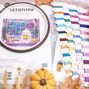 May include: A cross-stitch kit with a fall theme, featuring a ghost holding a coffee cup, a pumpkin, and a cityscape. The kit includes a wooden hoop, colorful embroidery floss, and a pattern. The text on the kit reads "LETISTITCH" and "Pumpkin Latte Nights".