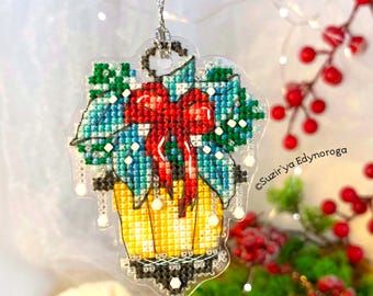 May include: A clear, cross-stitched Christmas ornament featuring a poinsettia design with a red bow. The ornament has a yellow pot, blue and green leaves, and white accents. It hangs from a silver string.