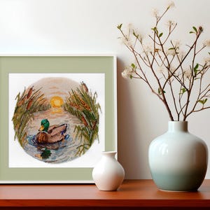 May include: A framed cross-stitch embroidery depicting a green-headed mallard duck swimming in a pond at sunset. The duck is surrounded by tall reeds and the setting sun is visible in the background.