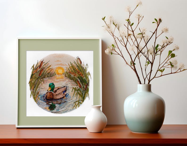 May include: A framed cross-stitch embroidery depicting a green-headed mallard duck swimming in a pond at sunset. The duck is surrounded by tall reeds and the setting sun is visible in the background.