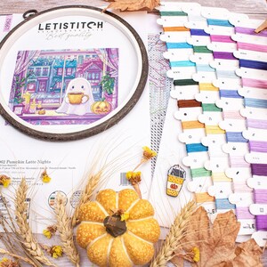 May include: A cross-stitch kit with a Halloween theme. The kit includes a wooden embroidery hoop with a stitched design of a ghost drinking coffee, a pumpkin, and colorful floss. Dried leaves and wheat stalks are arranged around the kit.