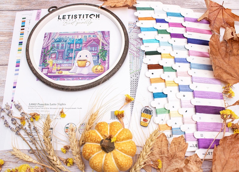 May include: A cross-stitch kit with a Halloween theme. The kit includes a wooden embroidery hoop with a stitched design of a ghost drinking coffee, a pumpkin, and colorful floss. Dried leaves and wheat stalks are arranged around the kit.