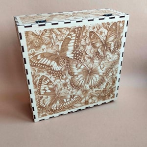 Puede incluir: Una caja de madera marrón claro con un diseño de mariposas y flores grabado con láser. Los bordes son de un color más claro con un borde decorativo. El diseño detallado es una característica destacada.