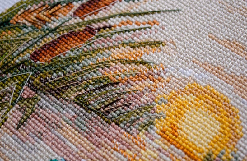 May include: Close-up of a cross-stitch embroidery with a detailed design of green leaves and yellow flowers. The fabric is white with a subtle texture.
