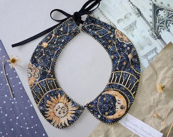 May include: A beaded collar necklace with a celestial design. The necklace features a sun, moon, and stars in gold and blue beads on a black fabric base.