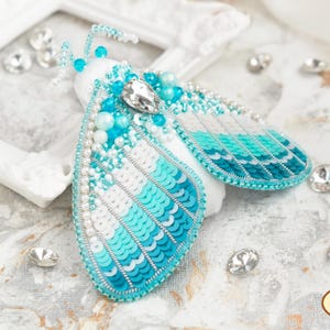 May include: A turquoise and white beaded moth brooch with a clear rhinestone accent. The moth is made with sequins and beads and has a gradient of color from white to turquoise.