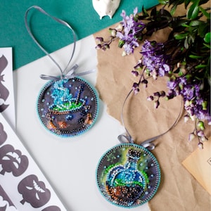May include: Two round, beaded ornaments with a Halloween theme. One ornament features a cauldron with blue smoke and the other features a potion bottle with a cork stopper. Both ornaments have a blue and green color scheme.