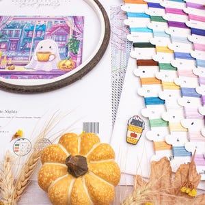 May include: A cross-stitch kit with a fall theme, featuring a ghost holding a mug, a pumpkin, and a cityscape scene. The kit includes embroidery floss in various colors, a pumpkin-shaped decoration, and a coffee cup pin.