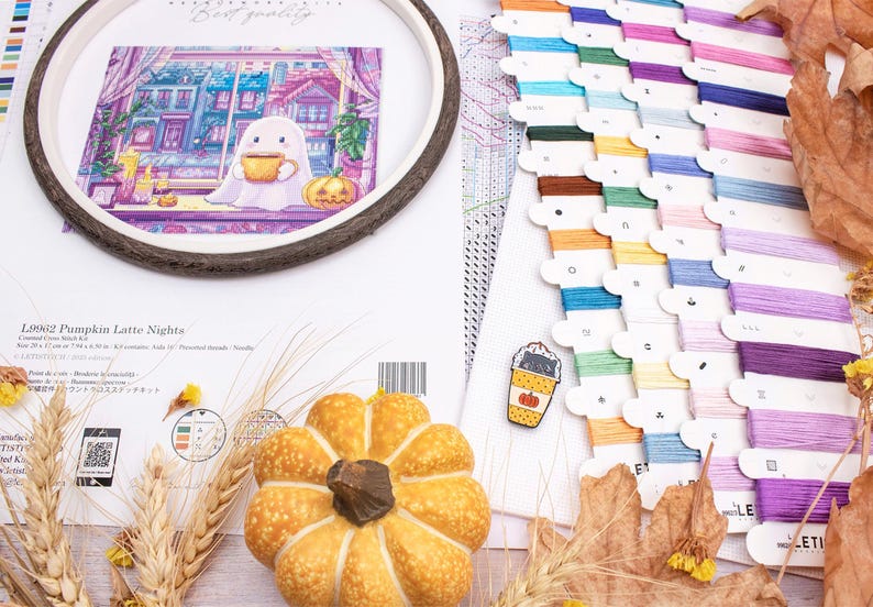 May include: A cross-stitch kit with a fall theme, featuring a ghost holding a mug, a pumpkin, and a cityscape scene. The kit includes embroidery floss in various colors, a pumpkin-shaped decoration, and a coffee cup pin.