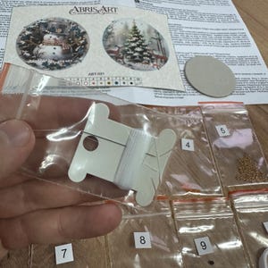 Peut inclure: Gros plan sur une bobine de fil blanc et neuf petits sacs contenant des perles et des paillettes. Les sacs sont num&eacute;rot&eacute;s de 4 &agrave; 9. La bobine de fil est dans un sac en plastique transparent. L'image est pour un kit de peinture au diamant.