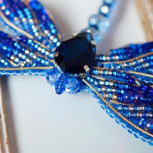 Puede incluir: Un broche de lib&eacute;lula azul hecho de cuentas y una gran piedra preciosa azul. El broche est&aacute; sobre un fondo blanco.