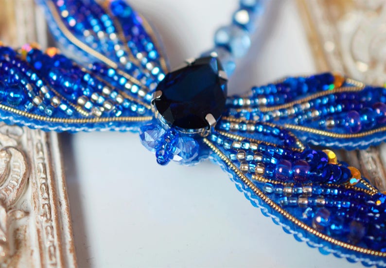 Puede incluir: Un broche de lib&eacute;lula azul hecho de cuentas y una gran piedra preciosa azul. El broche est&aacute; sobre un fondo blanco.