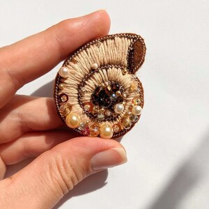 May include: A handcrafted seashell brooch with intricate embroidery in beige and brown tones. The design features a spiral pattern, embellished with pearls, beads, and crystals in shades of gold, brown, and amber. The brooch is a unique accessory.