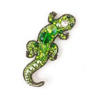 Op de afbeelding: Groene, met kralen bezette hagedisbroche met een gedetailleerd ontwerp. De broche heeft verschillende groentinten, waaronder facetkristallen en parels, omlijnd met zwarte kralen. Het lichaam van de hagedis is langwerpig, met kleine, met kralen bezette poten en een staart.