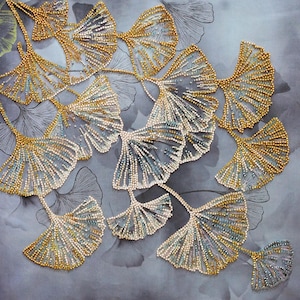 Könnte beinhalten: Nahaufnahme einer Perlen-Stickerei mit einem Cluster von Ginkgoblättern in Gold- und Blautönen. Die Blätter sind in einem kaskadierenden Muster auf einem grauen Stoffhintergrund angeordnet.