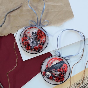 May include: Two round, beaded ornaments with a Halloween theme. One ornament features a skeleton with a skull and red berries. The other ornament features a black raven with a skull and red berries.