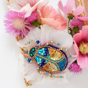 May include: A blue and gold jeweled beetle brooch with a beaded design, surrounded by pink and white flowers.