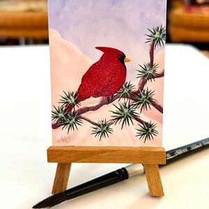 May include: A watercolour painting of a red cardinal perched on a branch of a snow-covered tree. The background is a light blue sky with white snow falling. The painting is signed "HC" in the bottom left corner.