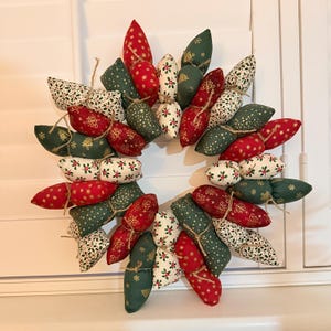 May include: A festive Christmas wreath composed of fabric pods in red, green, and white. The pods are adorned with gold stars, holly, and other holiday patterns. The wreath is tied together with twine and is displayed against a white shutter background.