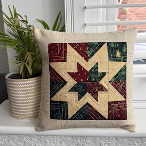 Puede incluir: Cojín decorativo cuadrado con un diseño de estrella acolchada. La estrella está compuesta por cuadrados de tela rojos, verdes y dorados. El cojín tiene un borde de color neutro y está colocado sobre una superficie blanca.