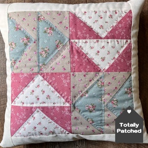 Op de afbeelding: Een wit kussen met een patchwork-ontwerp met roze, beige en blauwe bloemenstof. Het kussen is gestikt met wit garen en heeft een wit hart met de tekst "Totally Patched" in zwart.