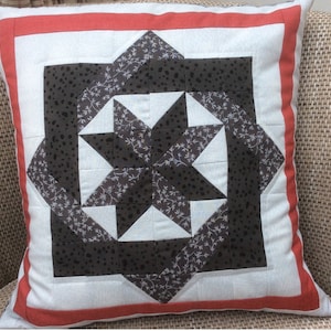 Puede incluir: Una almohada blanca con un borde rojo y un patrón geométrico marrón y blanco en el centro. El patrón es una estrella con un fondo blanco y acentos marrones.