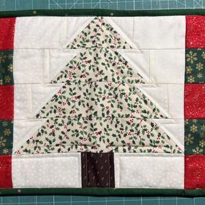 May include: A rectangular Christmas-themed quilted placemat. The center features a Christmas tree design with holly leaves and red berries. The sides have red and green squares with gold snowflakes. The border is green.