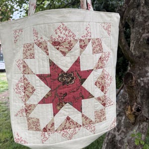 Puede incluir: Un bolso tote acolchado con un patrón de estrella central en tela floral de color rojo apagado y beige. El bolso está confeccionado en tela color blanco roto con líneas de costura verticales y horizontales que forman una cuadrícula.