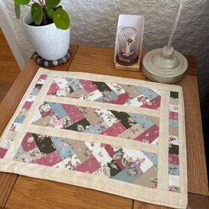 May include: A rectangular quilted placemat with a floral pattern in pink, blue, and beige. The placemat has a cream-colored border and is placed on a wooden table. A small potted plant and a decorative item are also visible.