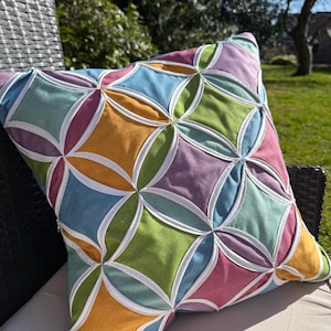 May include: A colorful geometric patterned pillow with a teal, green, purple, and orange design. The pillow is made of fabric and has a white border.