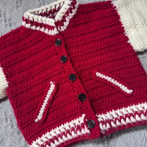May include: Red and white crocheted cardigan with a button closure and two pockets. The cardigan has a varsity jacket style with white trim.